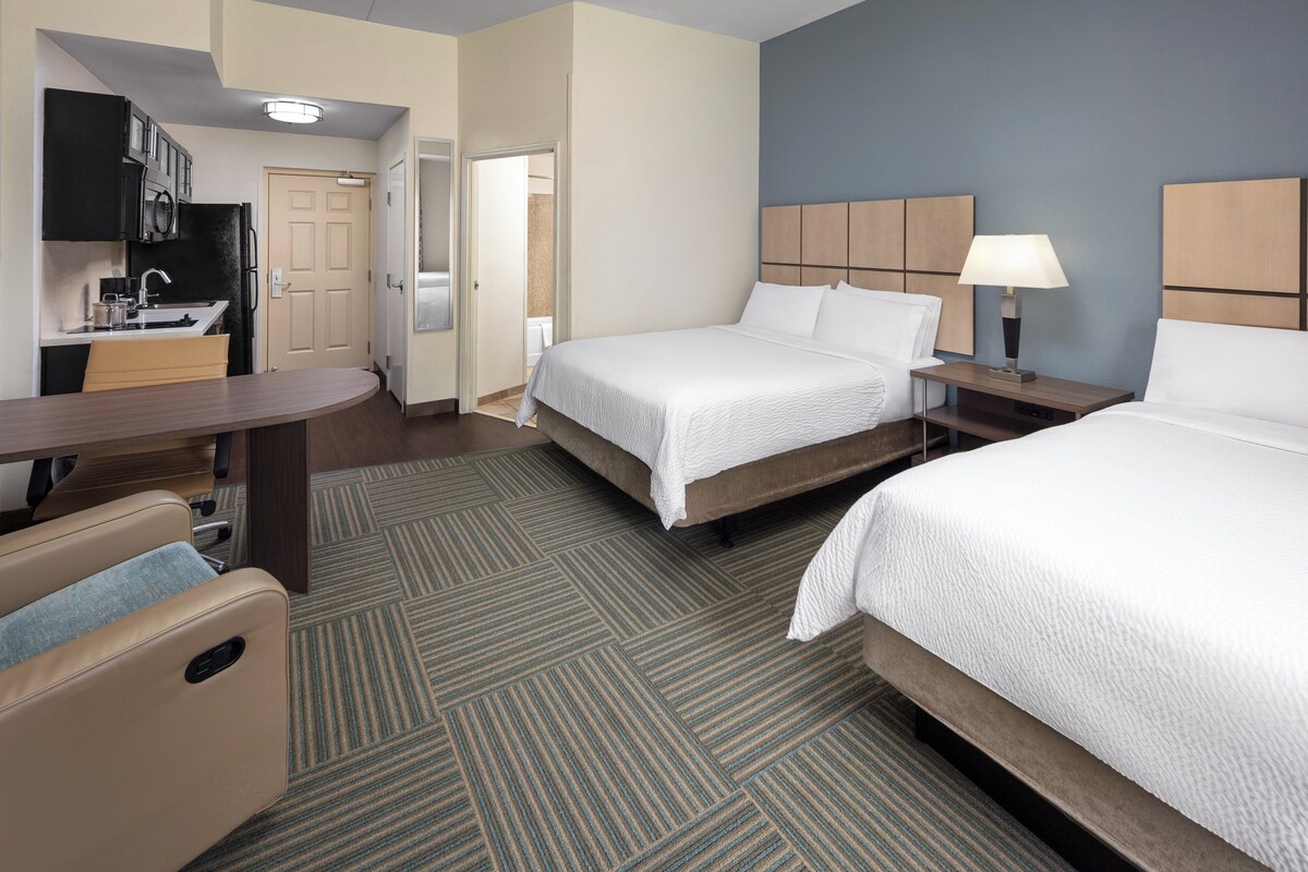 A hotel room with a bed, bedside table, lamp, and a kitchenette area.