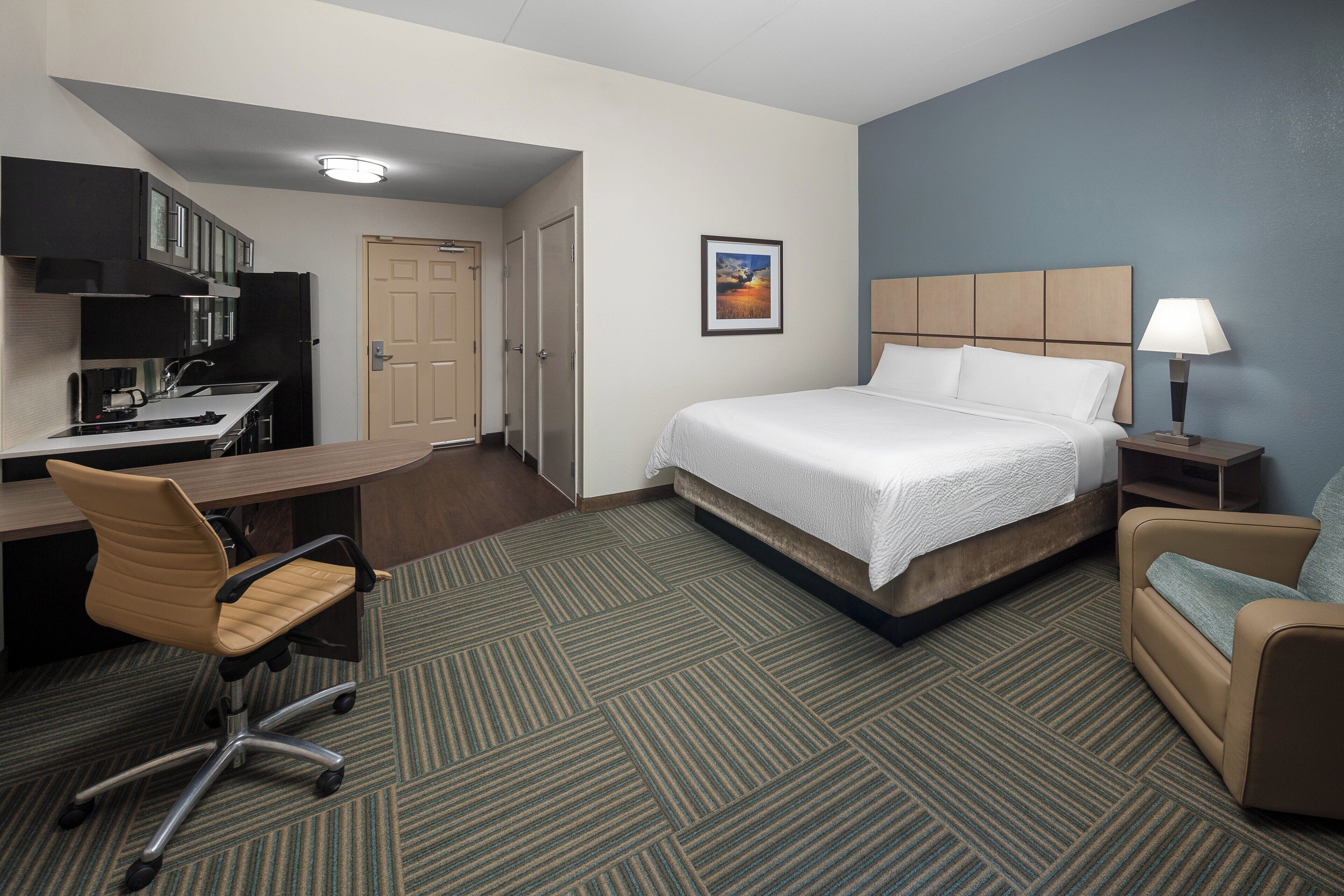 A hotel room with a bed, desk, chair, kitchenette, and a painting on the wall.