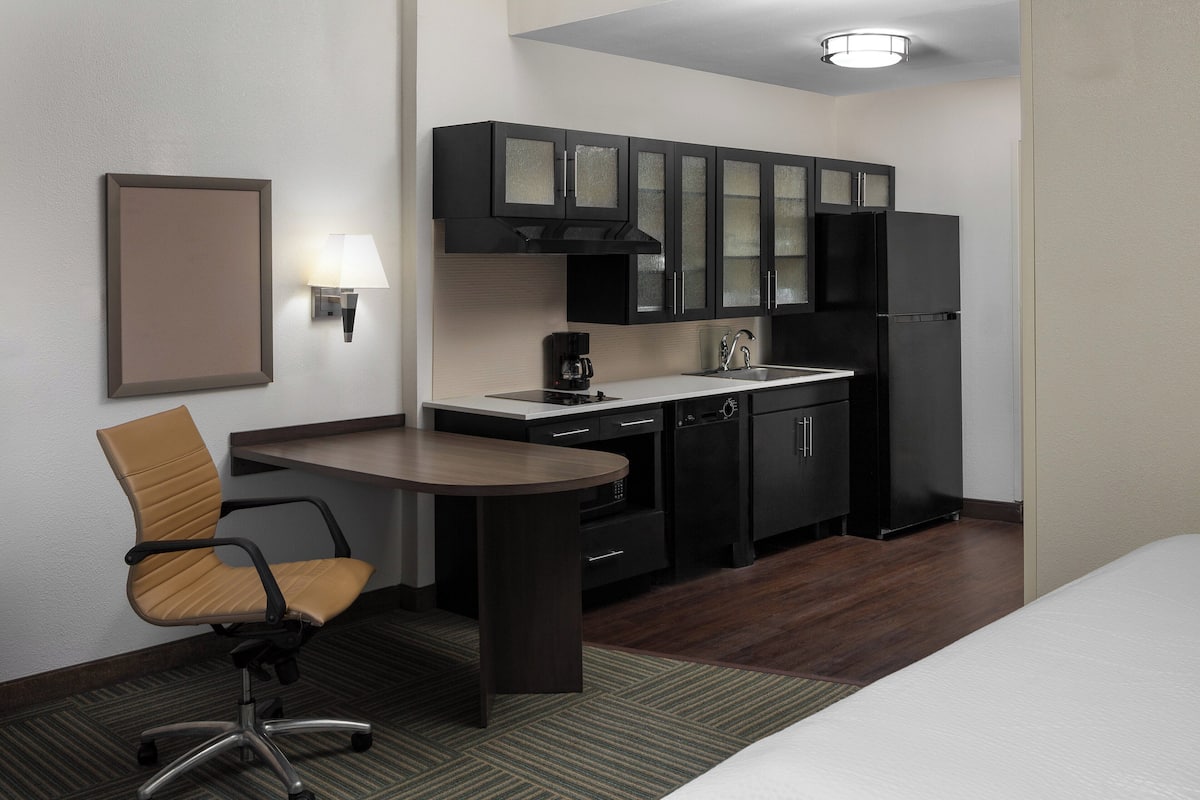 Una habitación de hotel compacta con kitchenette, una cama individual, una silla y una mesita.
