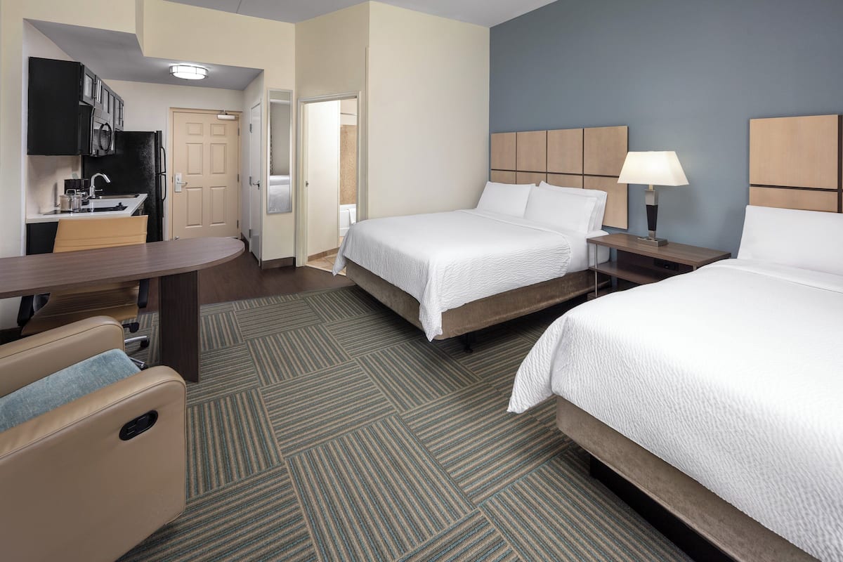 Una habitación de hotel con cama, mesita de noche, lámpara y una zona de kitchenette.