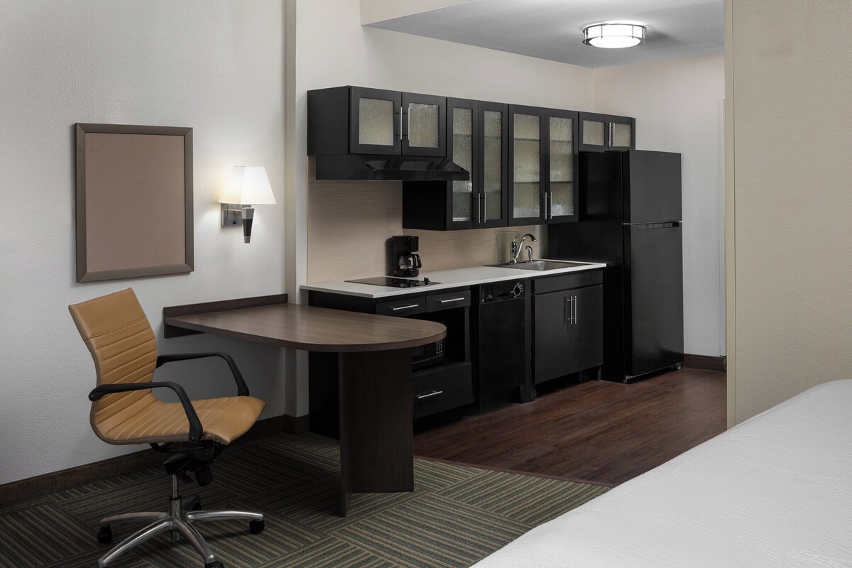 A compact hotel room with a kitchenette, a single bed, a chair, and a small table.