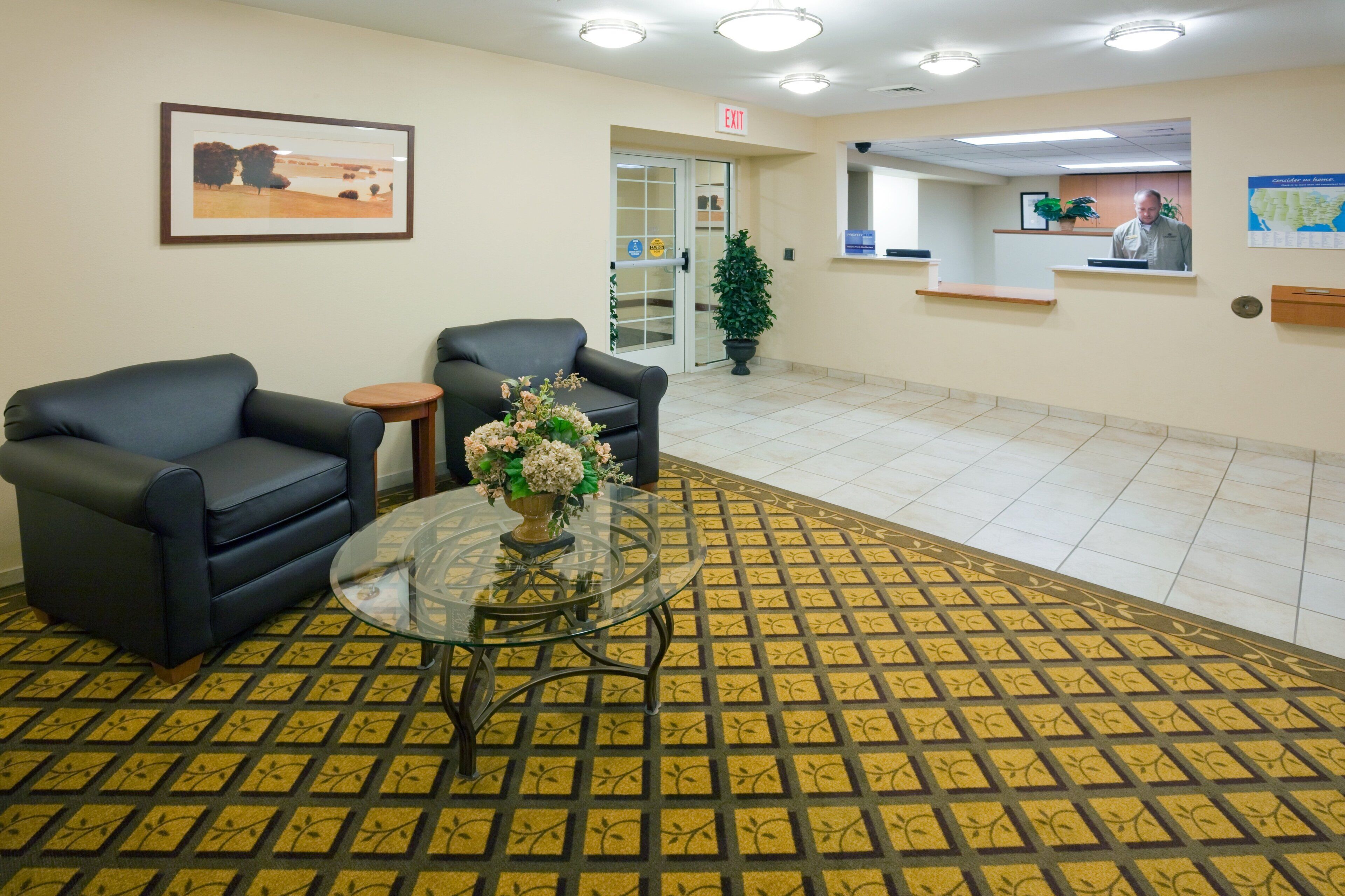 A reception area with two black sofas, a glass coffee table with a floral centerpiece, a patterned carpet, and a reception desk with a person behind it.