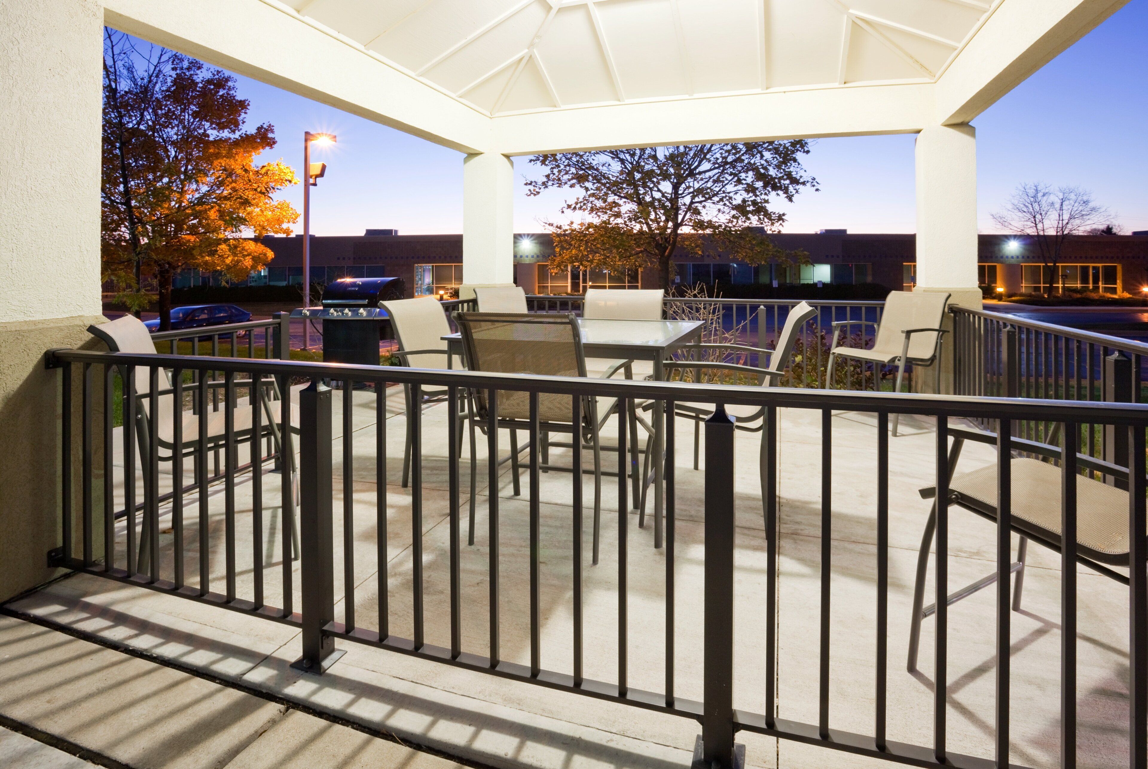 An outdoor patio area with tables and chairs, a railing, and a view of a parking lot and buildings in the background.