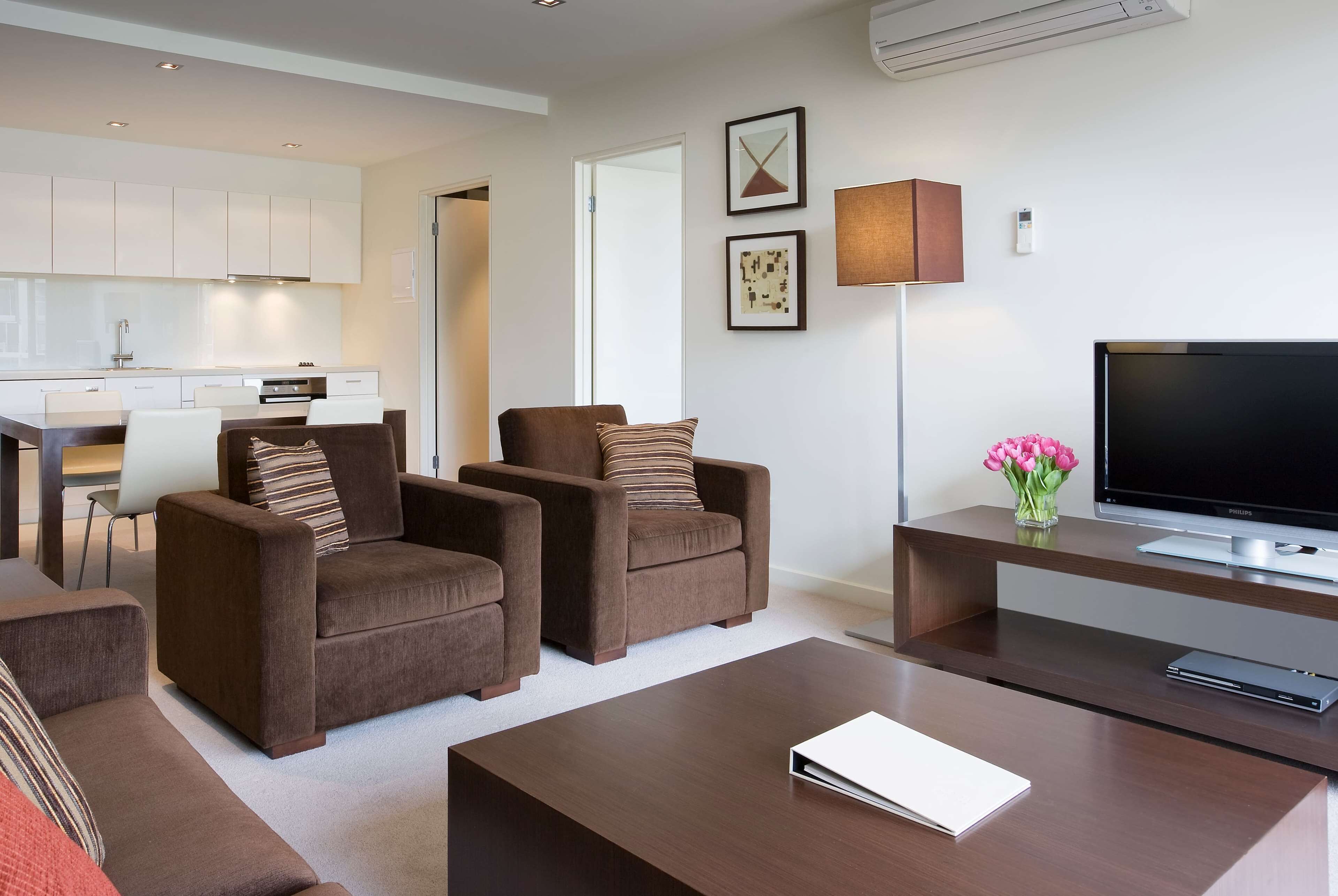 A modern living room with a flat-screen TV, two brown armchairs, a coffee table, and a dining area with a kitchen in the background.