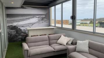 Un soggiorno moderno con un divano angolare grigio, un tavolino da caffè e un'ampia finestra con vista sulla spiaggia e sull'oceano.