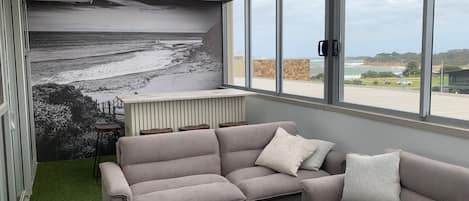 A modern living room with a grey sectional sofa, a coffee table, and a large window offering a view of the beach and ocean.
