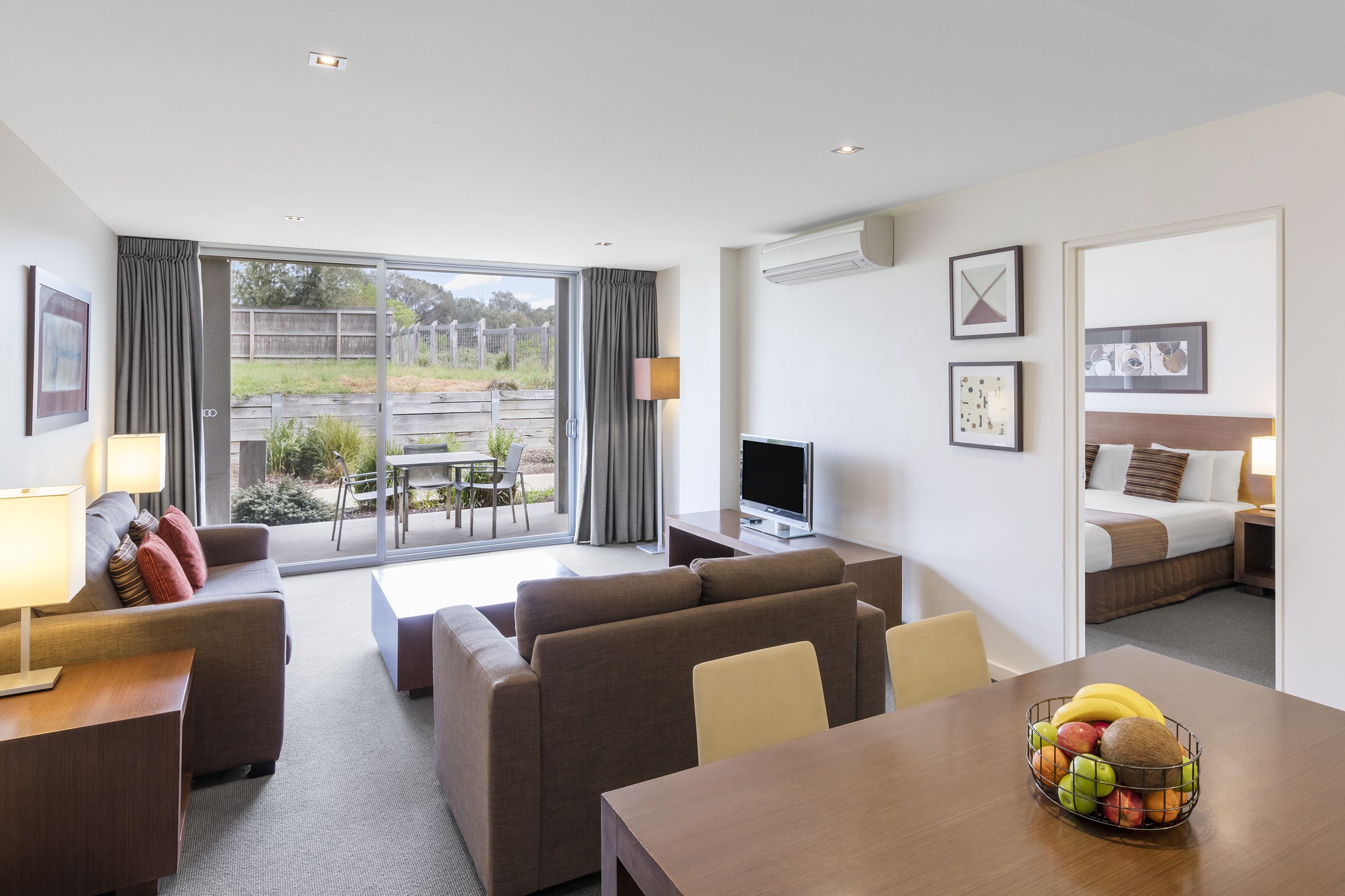 Una moderna camera d'albergo con un'ampia porta-finestra scorrevole verso l'esterno, una TV a schermo piatto, un'area pranzo con un cestino di frutta e un letto con testiera.