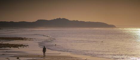Una playa con una persona caminando por la orilla, colinas lejanas y agua tranquila.