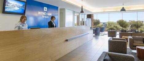 Zona de recepción con un mostrador de madera, dos recepcionistas, un gran letrero azul con el texto 'WYNDHAM Torquay', y vistas del exterior a través de amplias ventanas.
