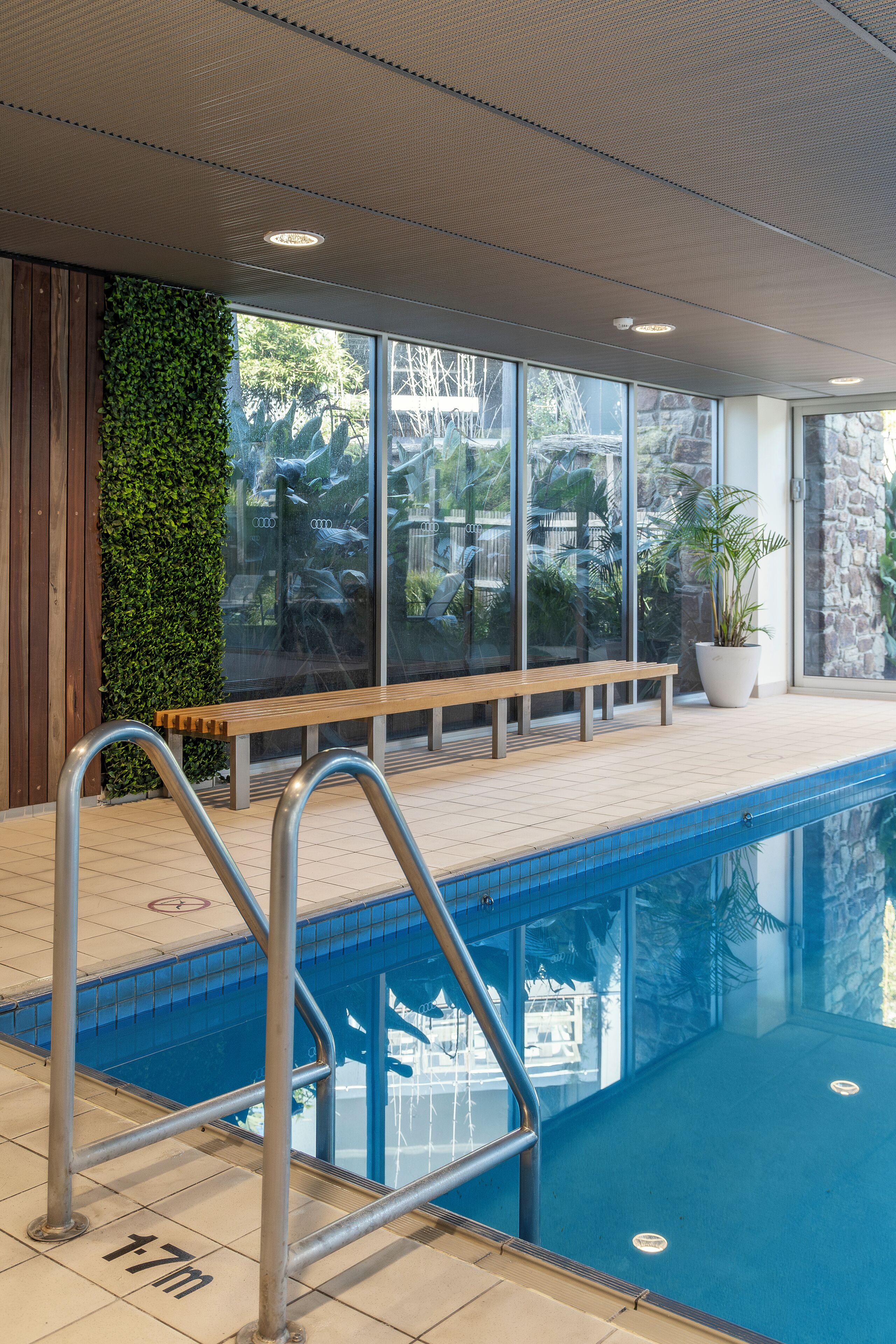 An indoor swimming pool with a wooden bench, metal pool ladder, and a green wall.