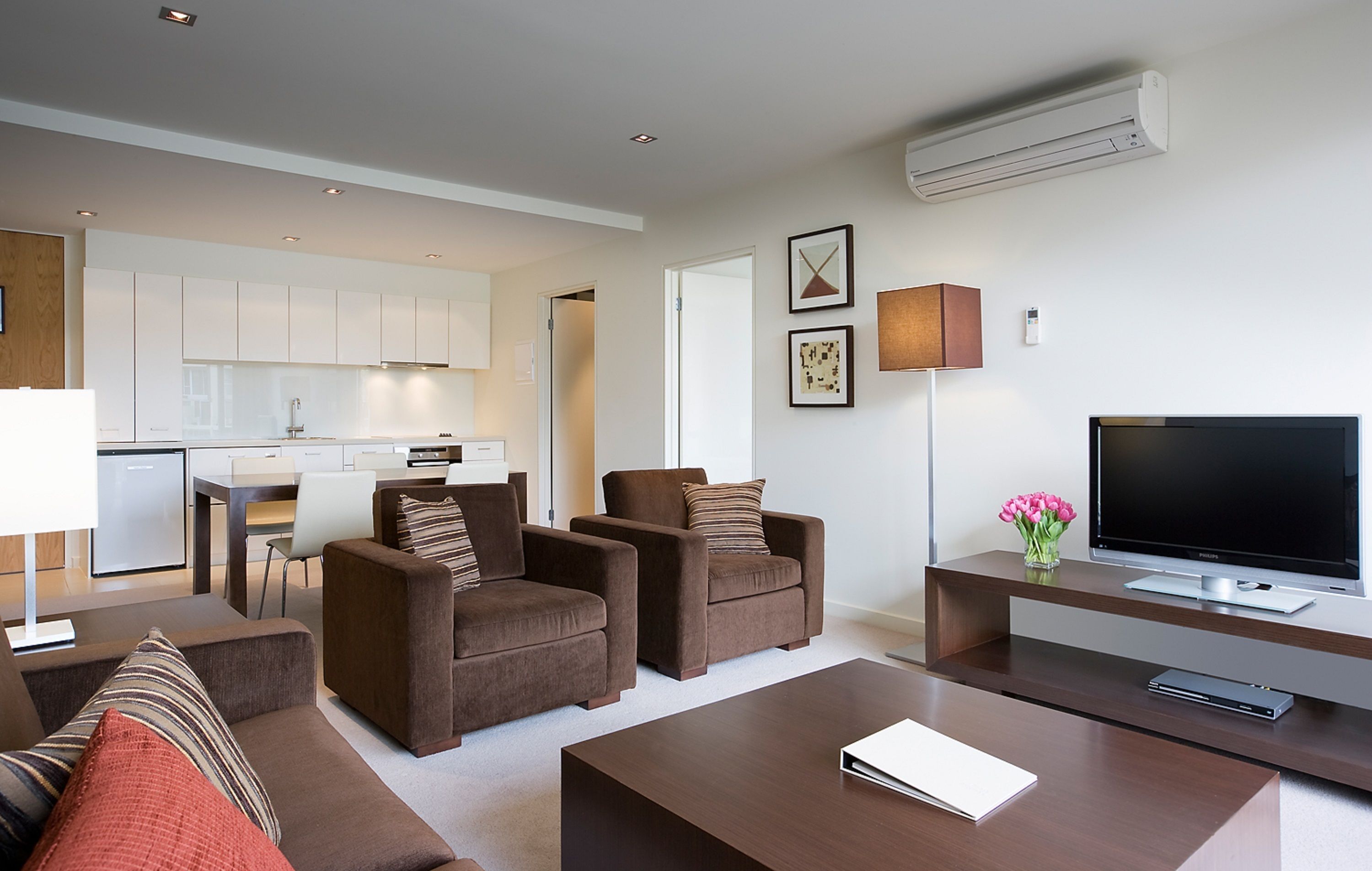 A modern living room with a flat-screen TV, brown sofas, a coffee table, and a kitchen in the background.