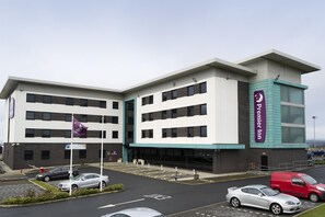 Et hotel med flere etager, et tydeligt 'Premier Inn' skilt, en parkeringsplads med flere biler og et flag på en flagstang.