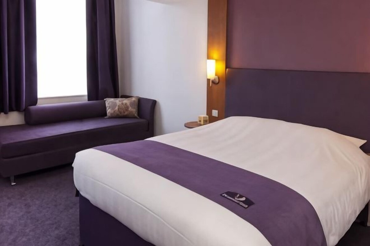 A hotel room with a bed, a sofa, a nightstand, and a wall-mounted light.