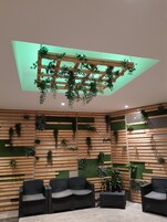Espaço interior moderno com ripas de madeira e plantas verdes nas paredes, teto com treliça de madeira e plantas penduradas, e mobiliário de vime preto.