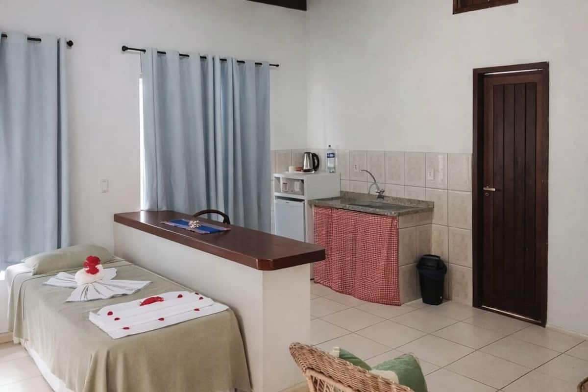 Una habitación con una cama, una kitchenette pequeña, una silla de mimbre y una puerta.