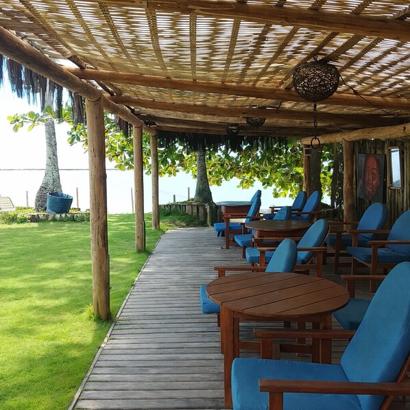 A covered outdoor seating area with wooden tables and blue chairs, a wooden deck, and a view of the sea. - Arraial Praia Hotel Pousada (Porto Seguro)