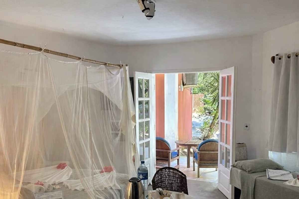 Una habitación con cama, una mesa con sillas, una mesita con tetera y tazas, y una ventana con cortinas.