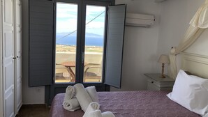 A bedroom with a bed, a window with open shutters, a view of the beach, and a nightstand with a lamp. - Aspa Villas (Santorini)