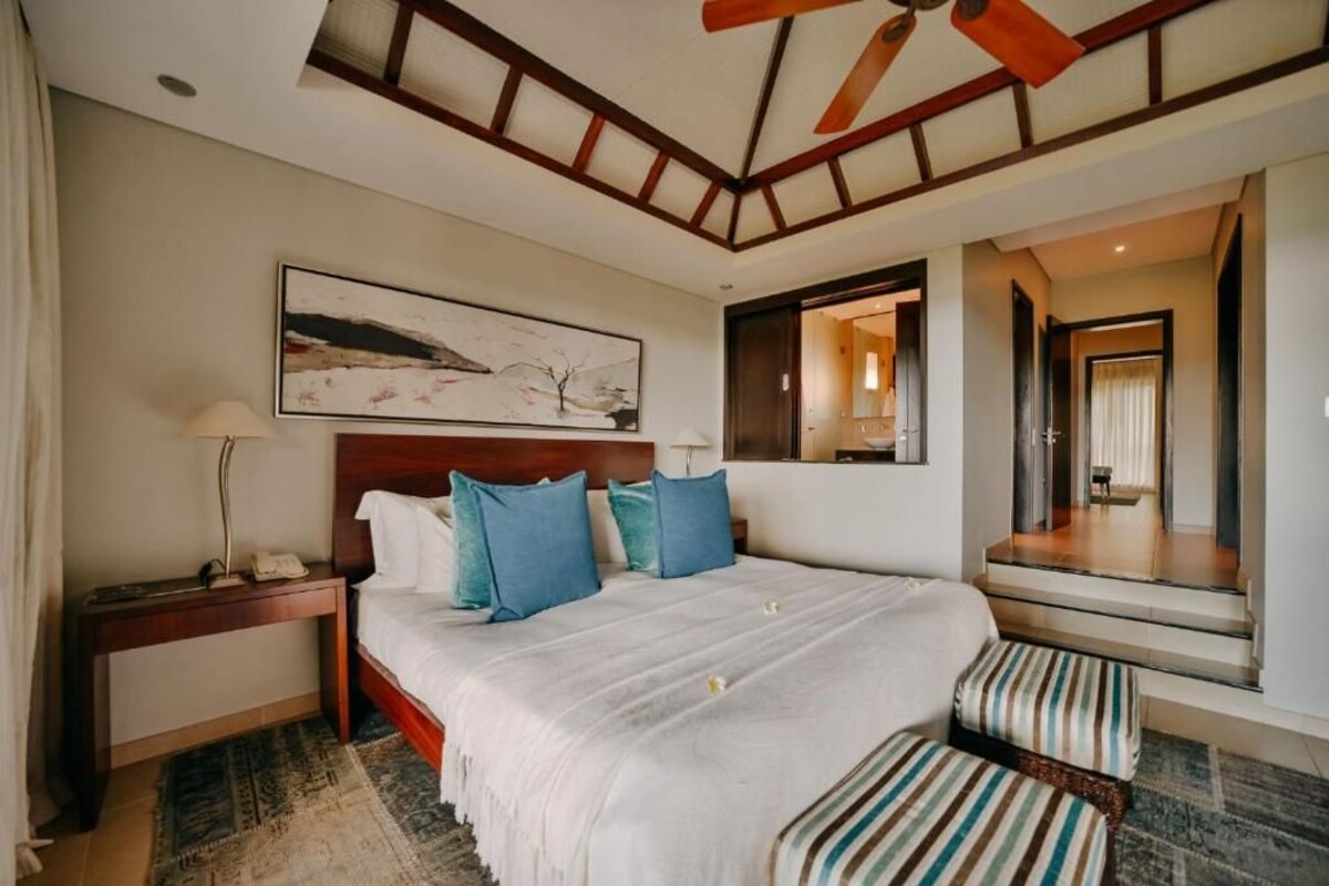 Una habitación de hotel con una cama grande, una mesita de noche, un escritorio, una silla, un ventilador de techo, un cuadro y un espejo.