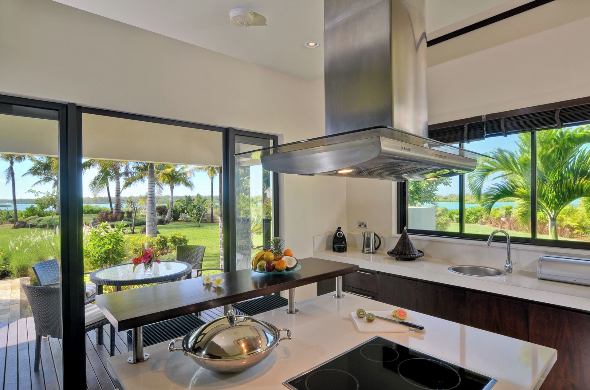 Une cuisine moderne dotée d’une grande fenêtre, offrant une vue sur un jardin tropical, et d’un coin repas avec des sièges d’extérieur.