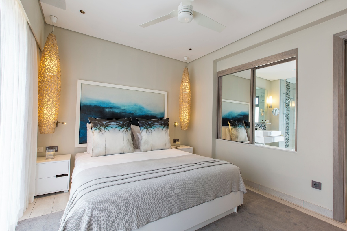 Un dormitorio moderno con una cama grande, ventilador de techo, espejo amplio y una obra de arte en la pared.
