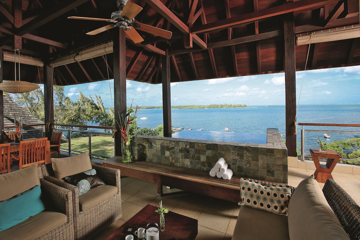 A covered outdoor seating area with wicker furniture, a stone fireplace, and a view of the sea.