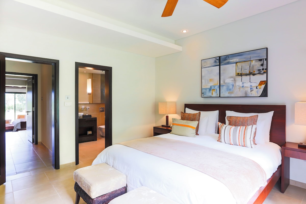 Una habitación de hotel con una cama grande, una mesita de noche con lámpara, un cuadro en la pared y un baño visible a través de una puerta abierta.
