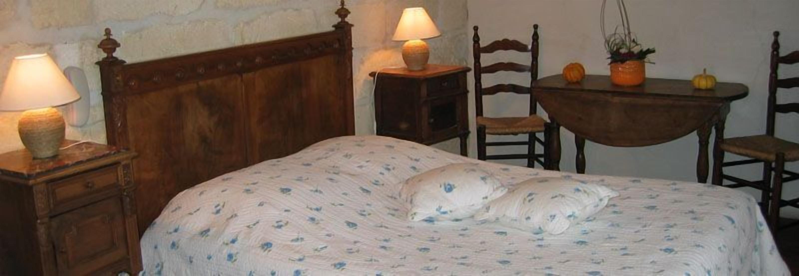 A bedroom with a wooden headboard, a bed with floral bedding, two bedside tables with lamps, a wooden chair, and a small table with a potted plant.