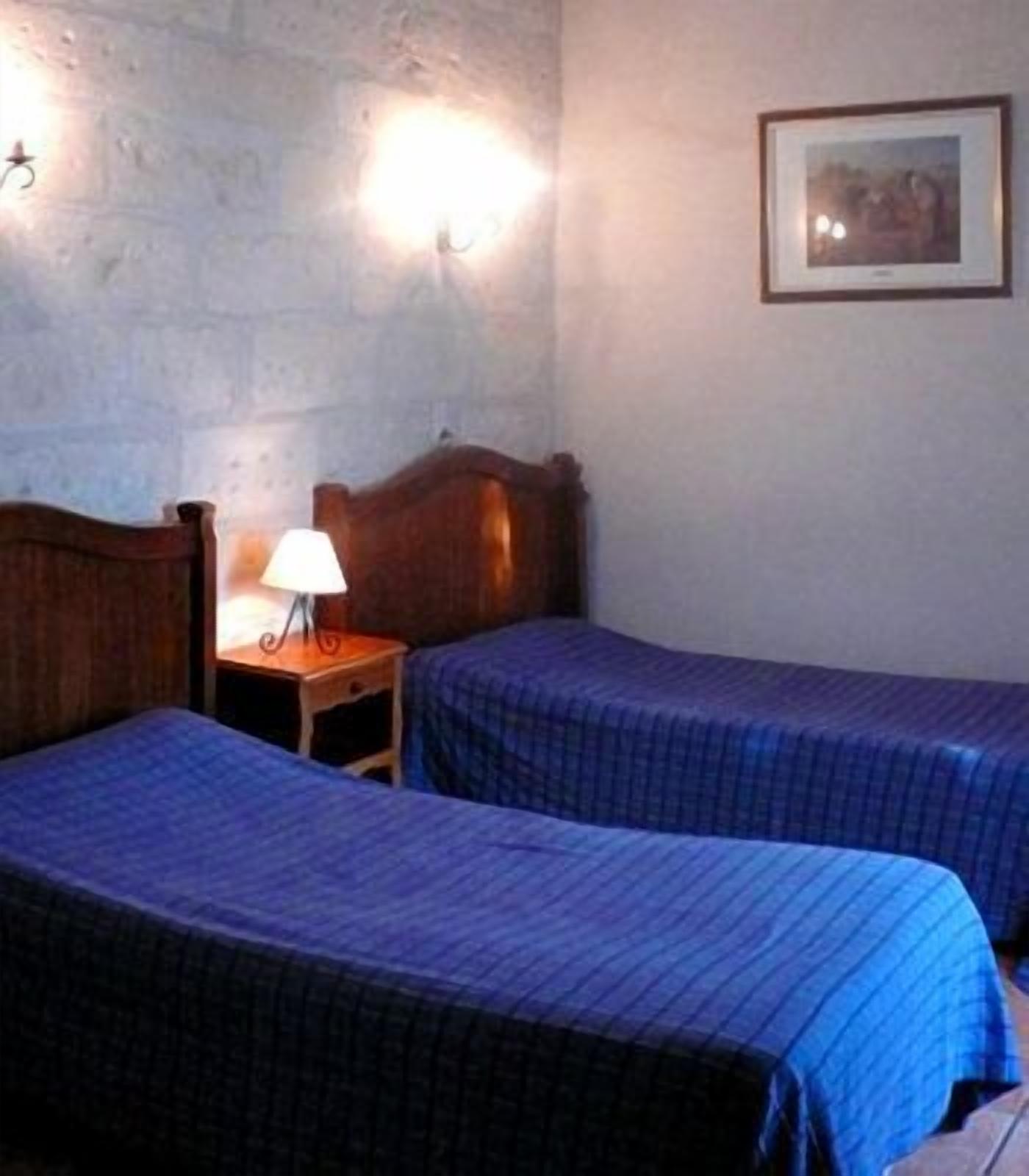 Una habitación con dos camas, cada una con cubrecama azul, una mesita de noche con lámpara y un cuadro en la pared.