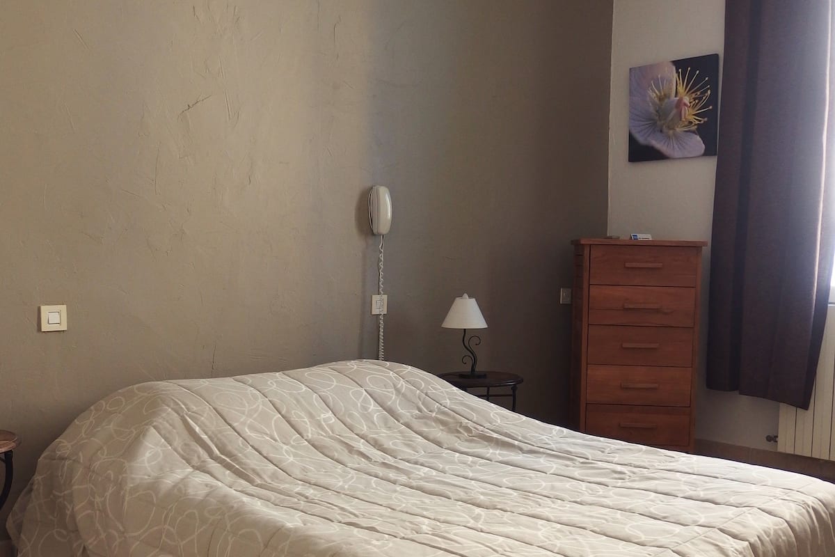 A bedroom with a large bed, a bedside table with a lamp, a dresser, and a wall-mounted telephone.