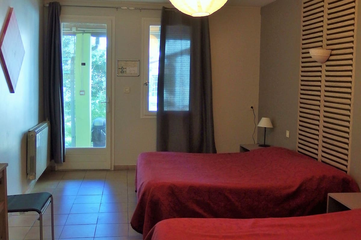 A hotel room with two beds, a radiator, a desk, a chair, a lamp, and a view of the outside through a door.
