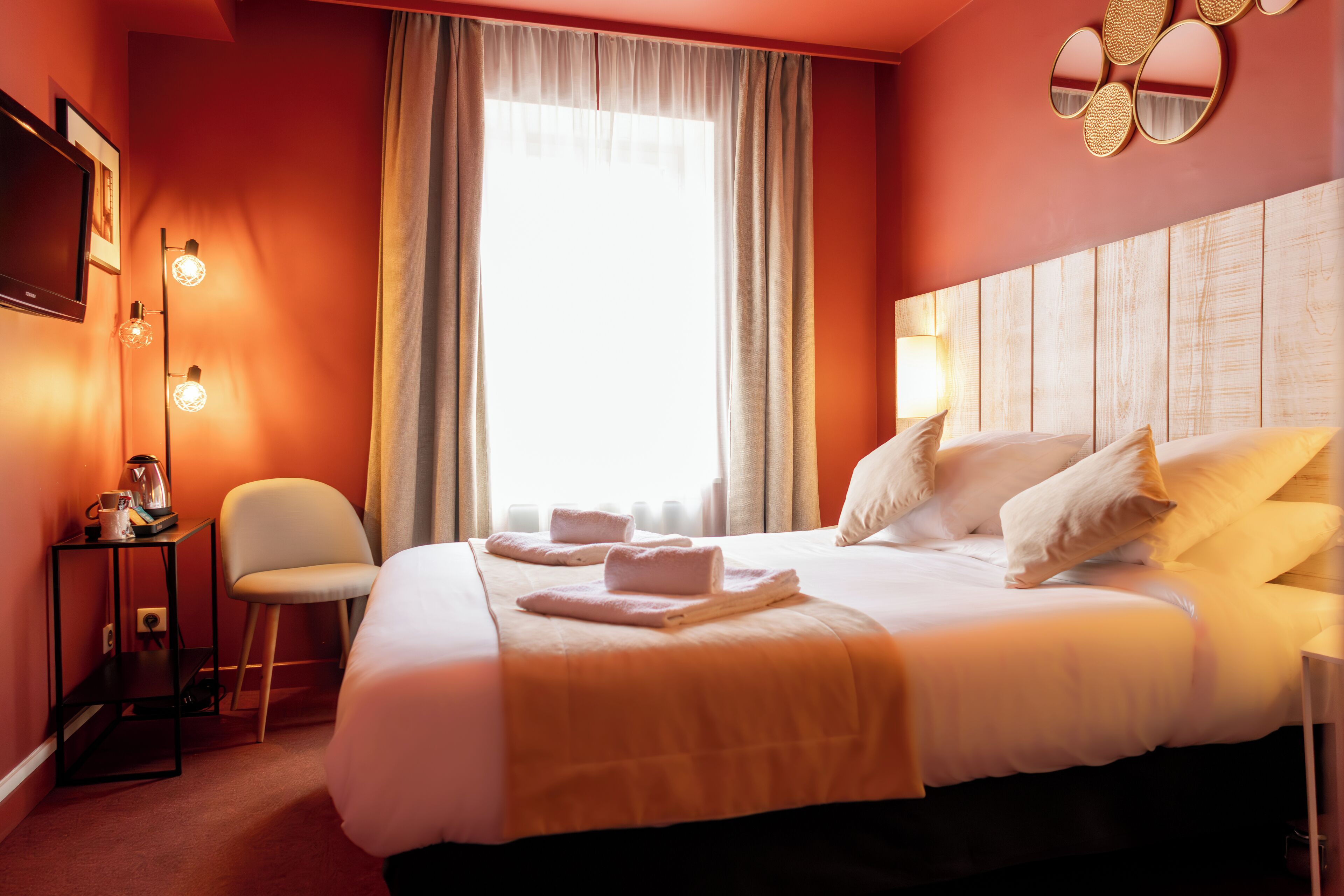 A hotel room with a large bed, a bedside table with a lamp, a chair, and a TV on the wall.