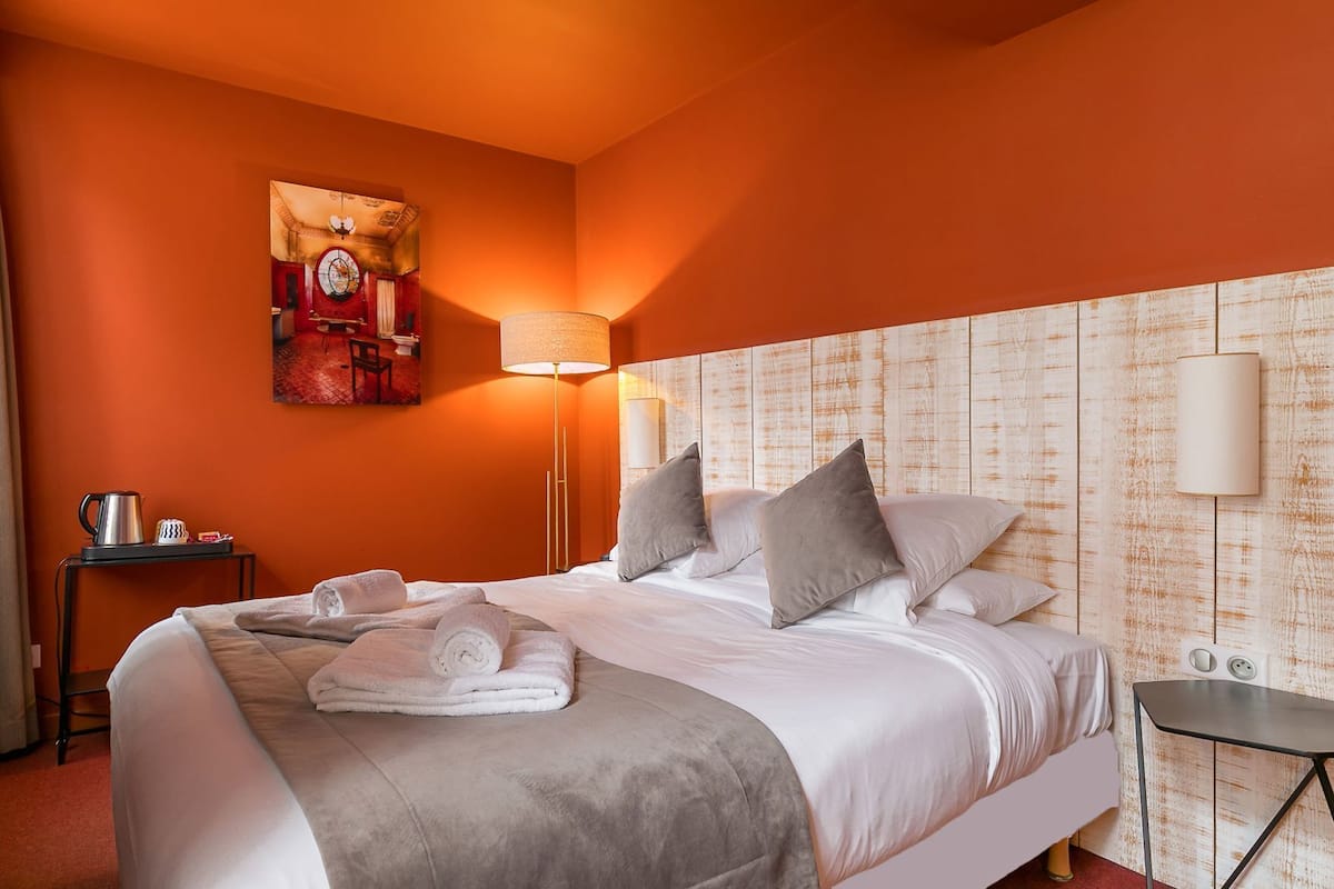 A hotel room with a large bed, a painting on the wall, a bedside table with a lamp, and a small table with a kettle.