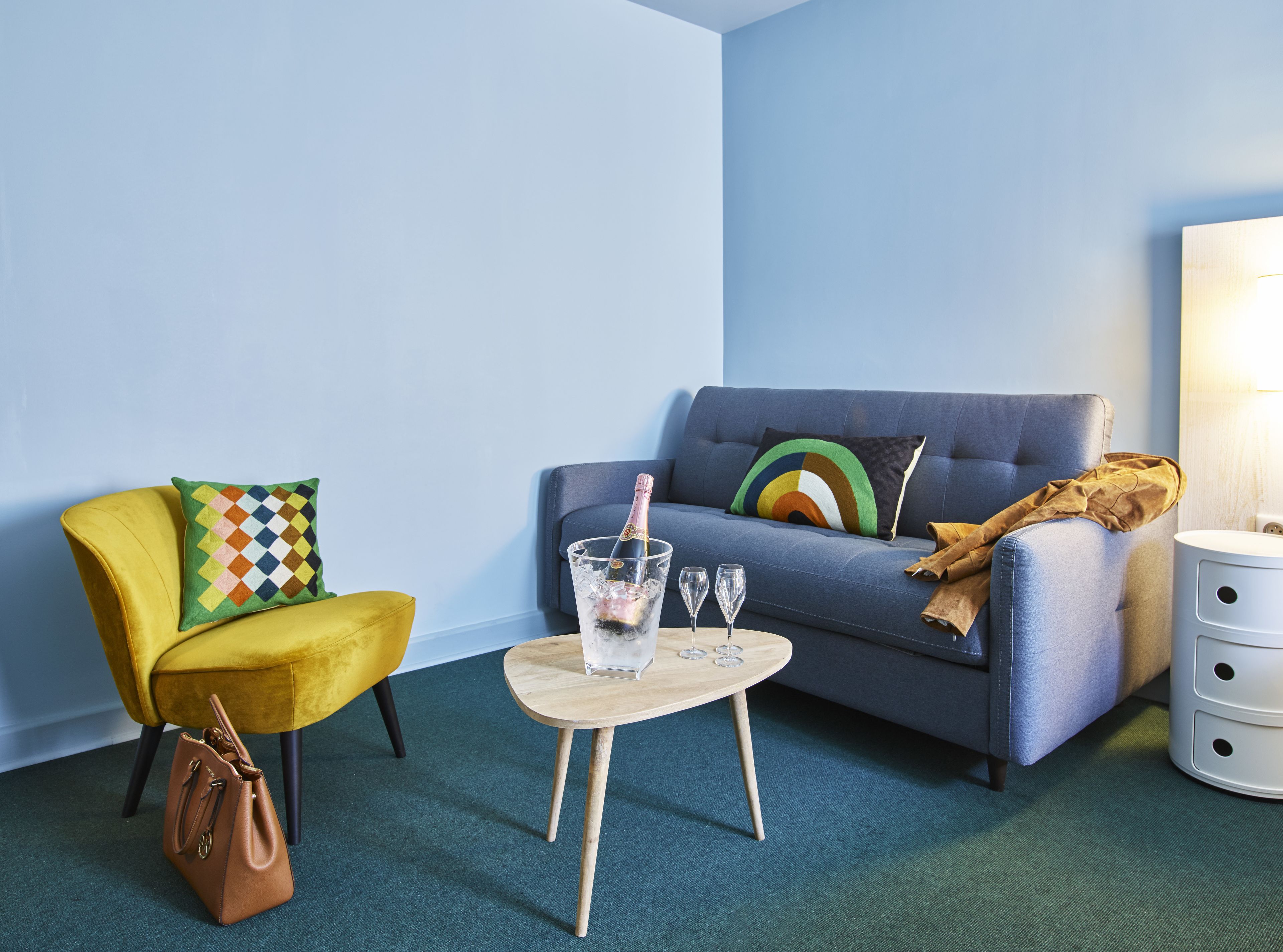A room with a yellow armchair, a blue sofa, a small wooden table with a champagne bottle and glasses, and a white bedside table with drawers.