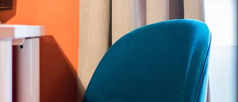 A teal chair with wooden legs, positioned against a wall with a beige curtain.
