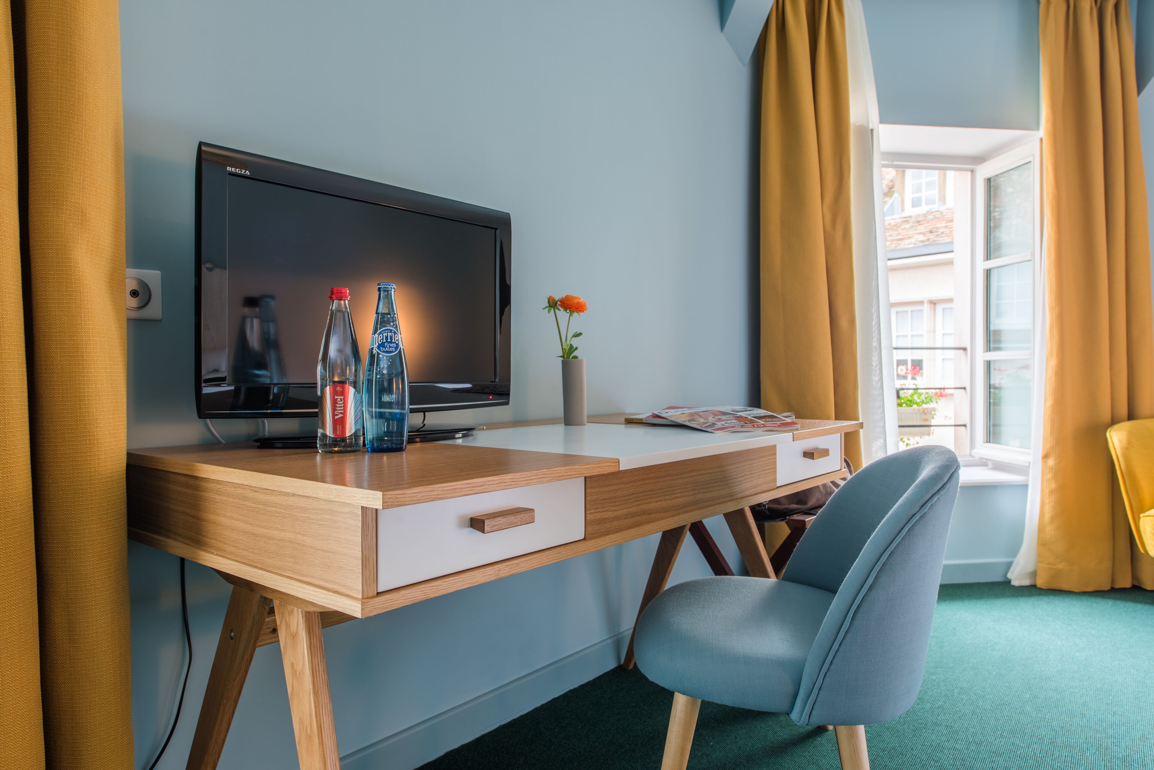 A hotel room with a wooden desk, a television, two bottles of water, a vase with flowers, and a chair.
