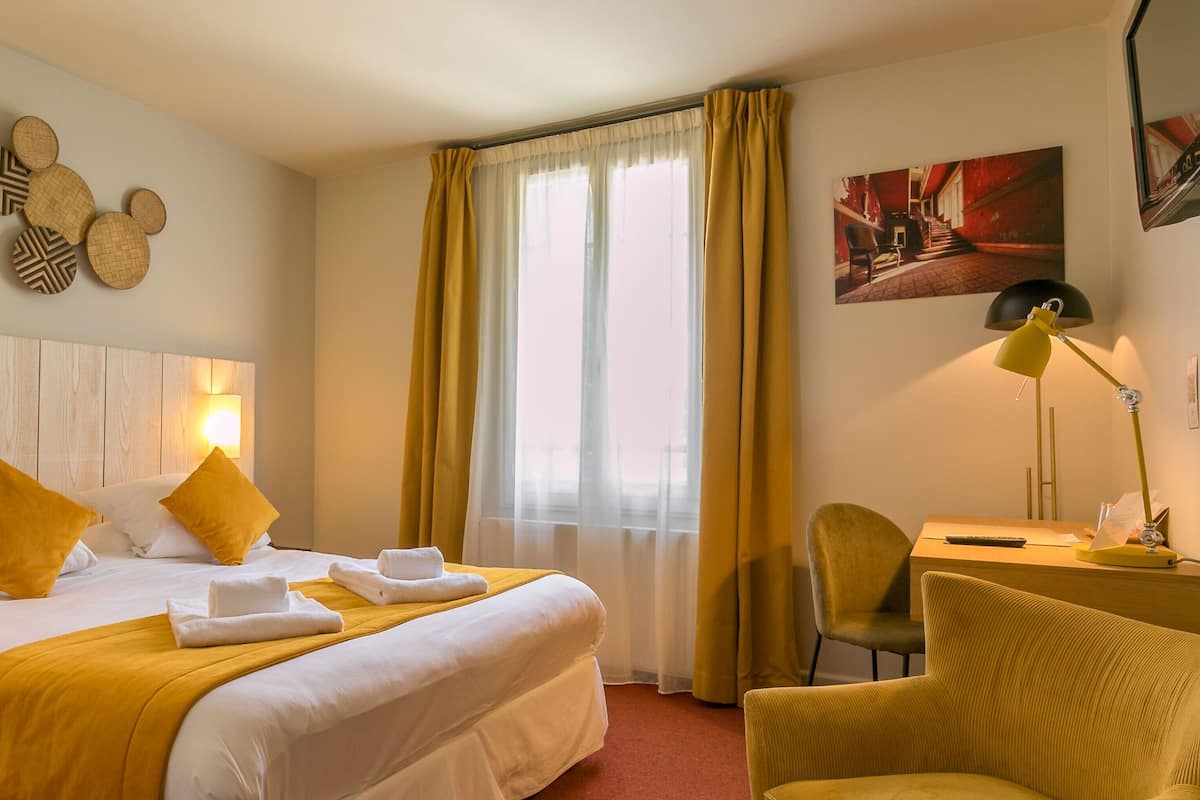 A hotel room with a wooden headboard, a bed with white linens and yellow pillows, a desk with a chair, a window with yellow curtains, and a painting on the wall.