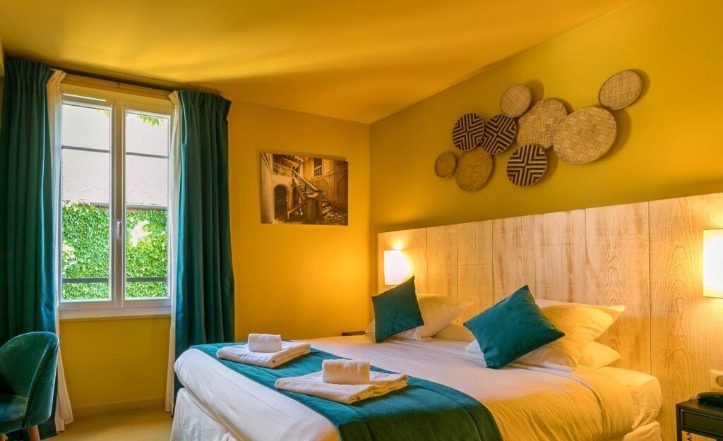 A hotel room with a bed, two pillows, a nightstand, a lamp, a window with curtains, and a wall decoration.