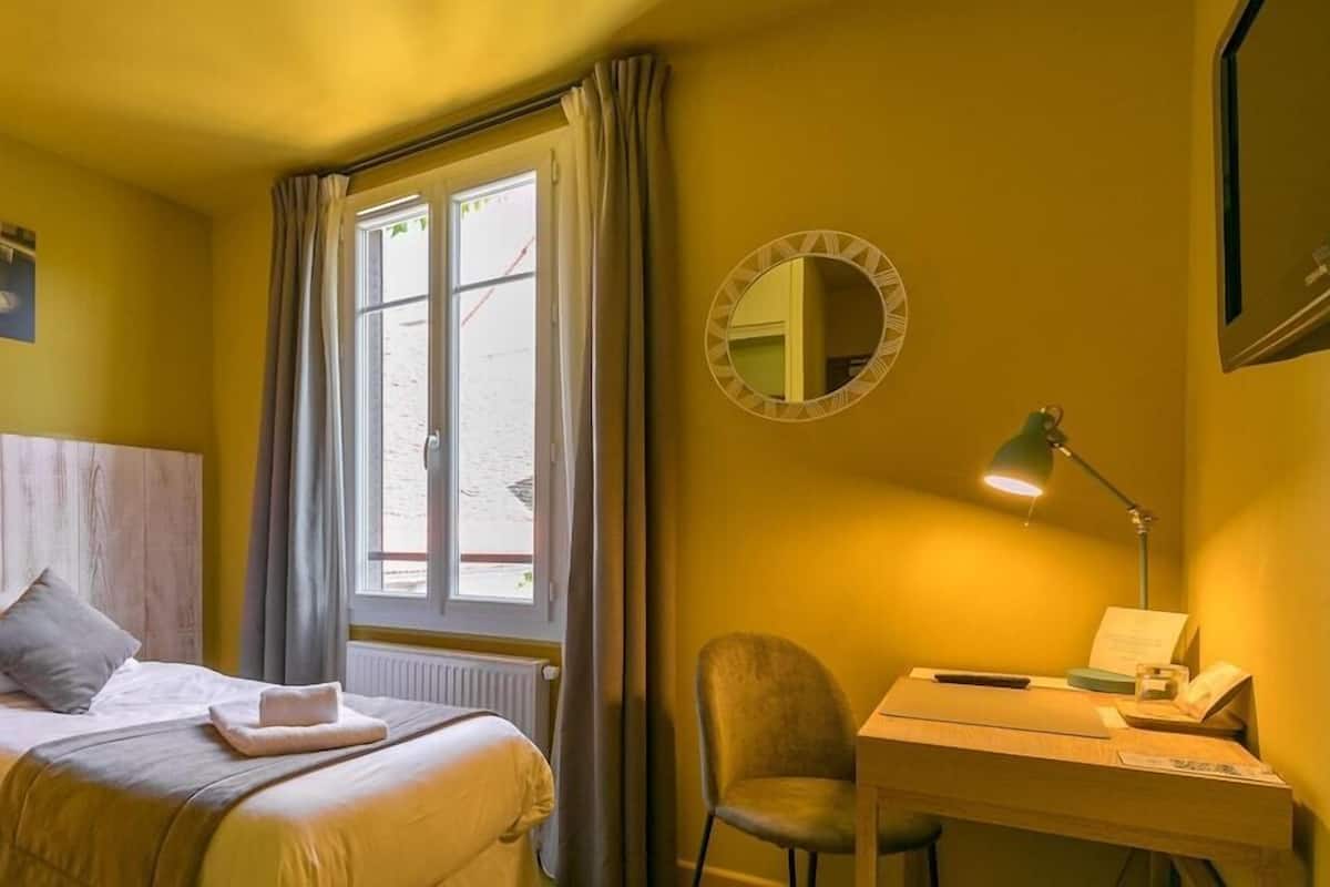 A hotel room with a bed, a desk, a chair, a window with curtains, a mirror, and a painting on the wall.