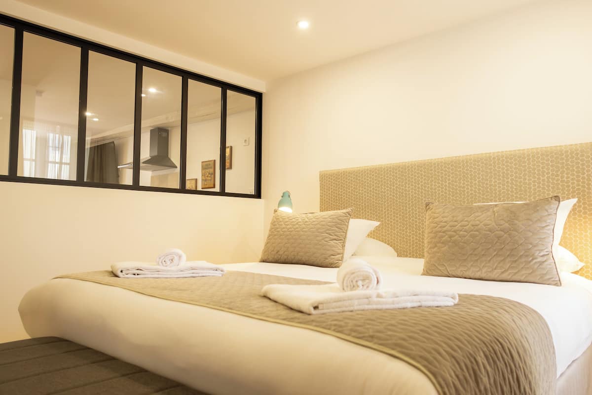 A hotel room with a large bed, two pillows, a headboard with a textured pattern, a mirror, and a window with curtains.