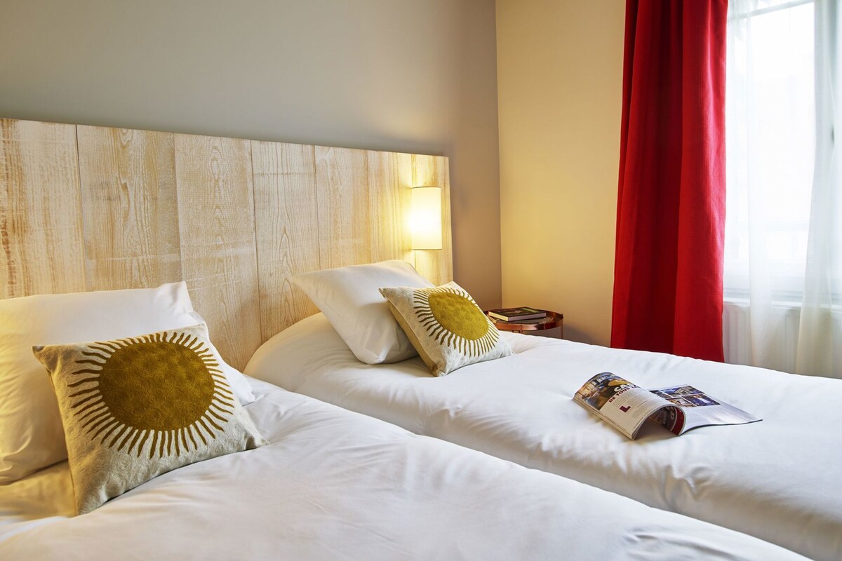 Een hotelkamer met twee bedden, een houten hoofdbord, een nachtkastje met een lamp en een raam met rode gordijnen.