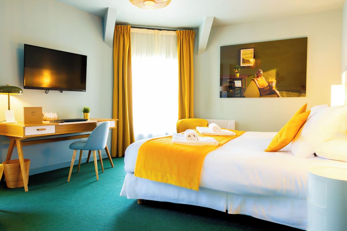 A hotel room with a bed, a desk, a chair, a TV, and a painting on the wall.