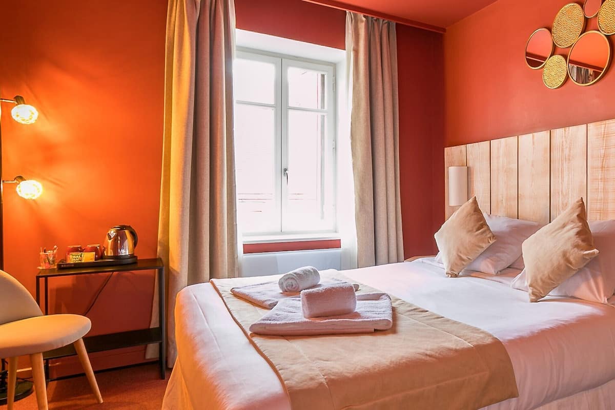A hotel room with a bed, a chair, a nightstand with a lamp, and a window with curtains.
