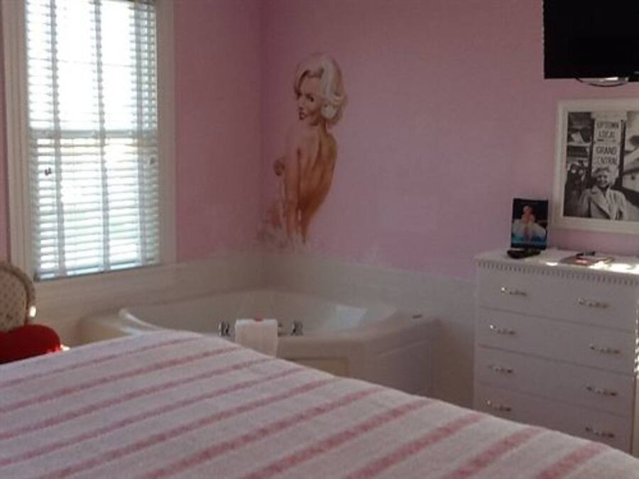 A bedroom with a large bed, a white dresser, a bathtub, and a pink wall with a mural.