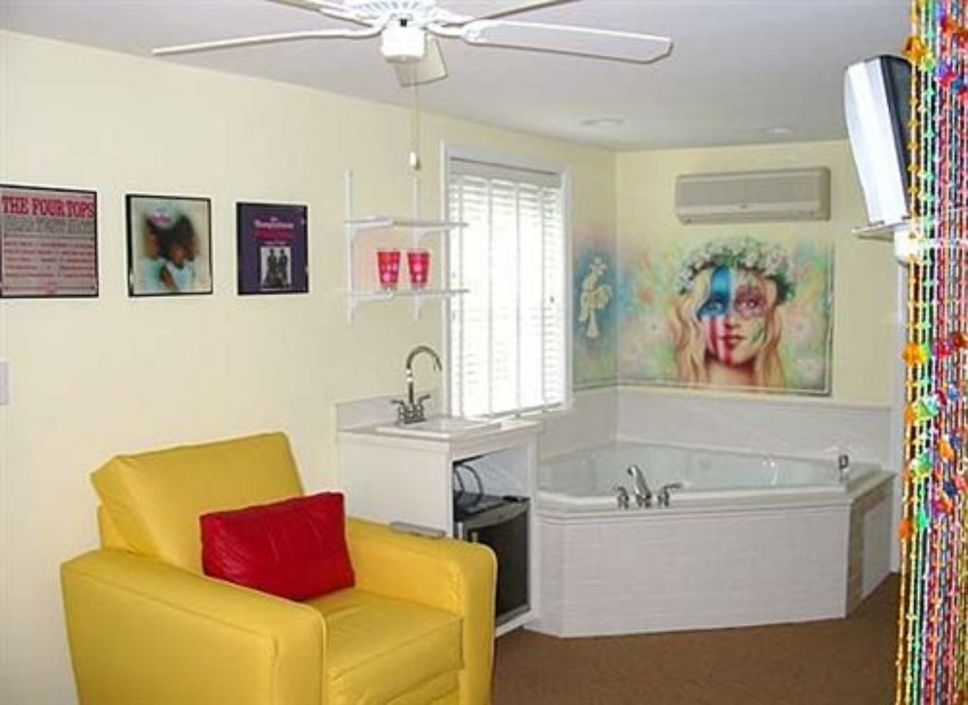 Una habitación con un sillón amarillo, una bañera blanca, un ventilador de techo y un cuadro en la pared.