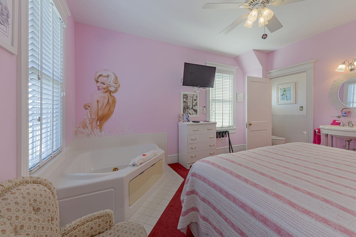 A bedroom with a large bed, a vanity with a mirror, a television mounted on the wall, and a window with blinds.