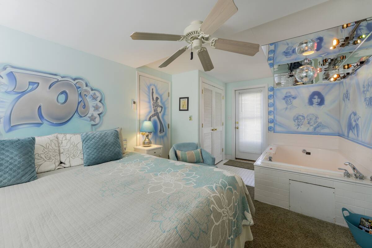 A bedroom with a large bed, a ceiling fan, a vanity with a mirror, and a bathtub.