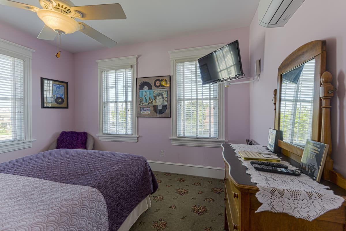 A bedroom with a bed, a dresser, a television mounted on the wall, and a window with blinds.
