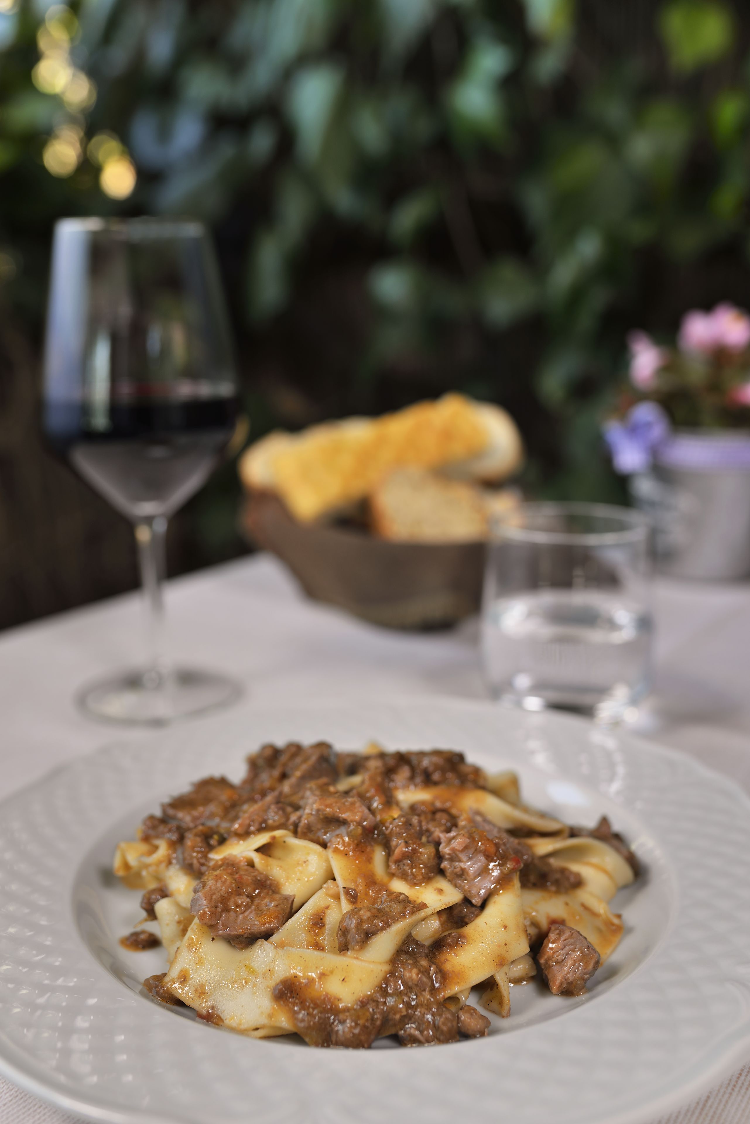 Un plato de pasta con salsa de carne, una copa de vino tinto y un bol de pan sobre una mesa.