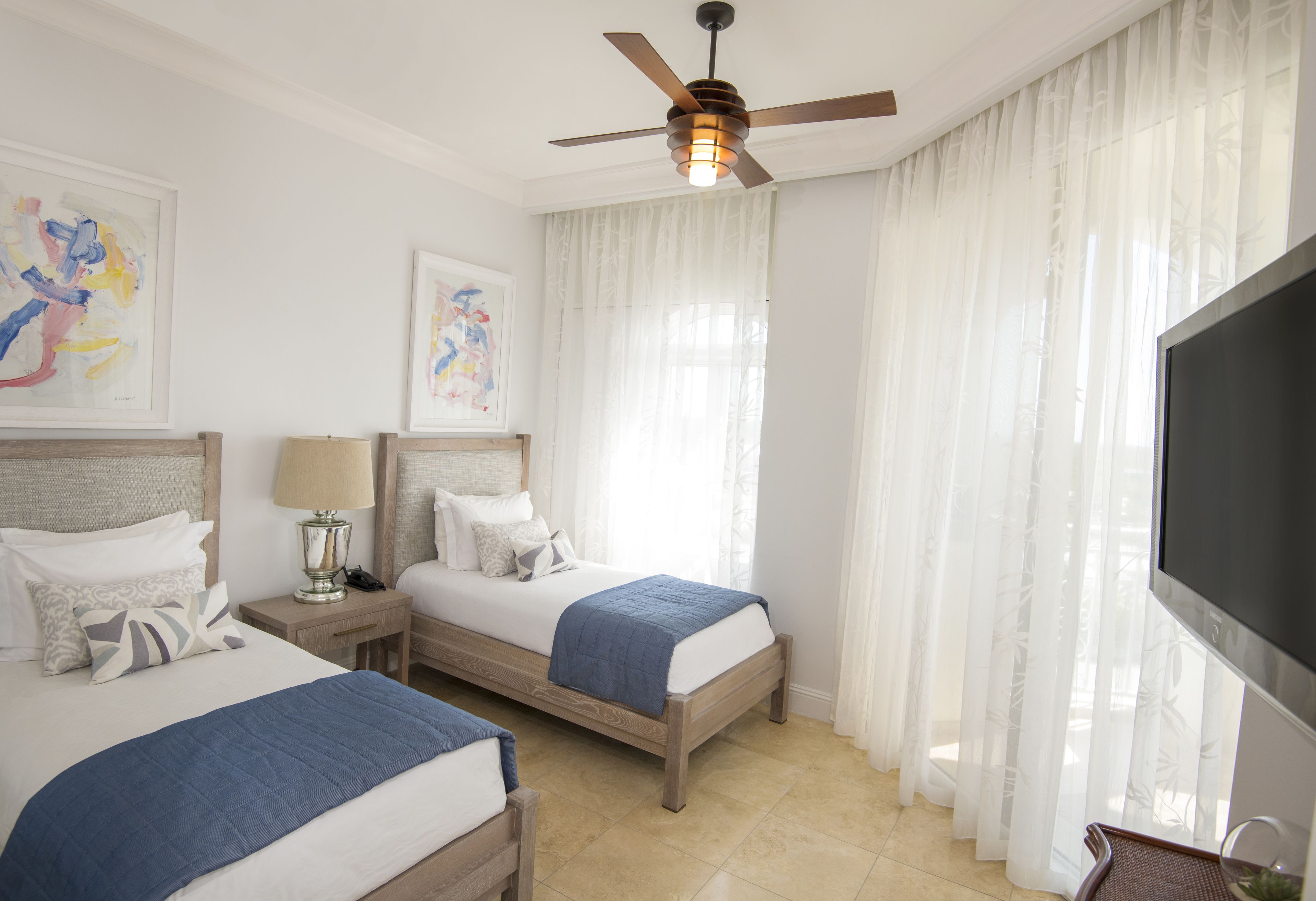 Quarto de hotel com duas camas, ventoinha no teto, televisão e obras de arte nas paredes.